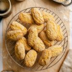 Overhead view of Italian sesame seed cookies (giuggiulena) piled on a round wire cooling rack over parchment paper, surrounded by two cups of black coffee, a bowl of sesame seeds, a small bowl of milk, and a linen napkin.
