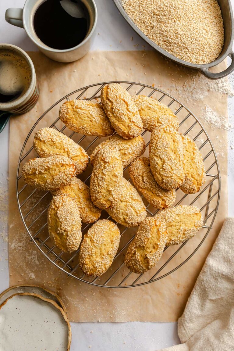 Overhead view of Italian sesame seed cookies (giuggiulena) piled on a round wire cooling rack over parchment paper, surrounded by two cups of black coffee, a bowl of sesame seeds, a small bowl of milk, and a linen napkin.