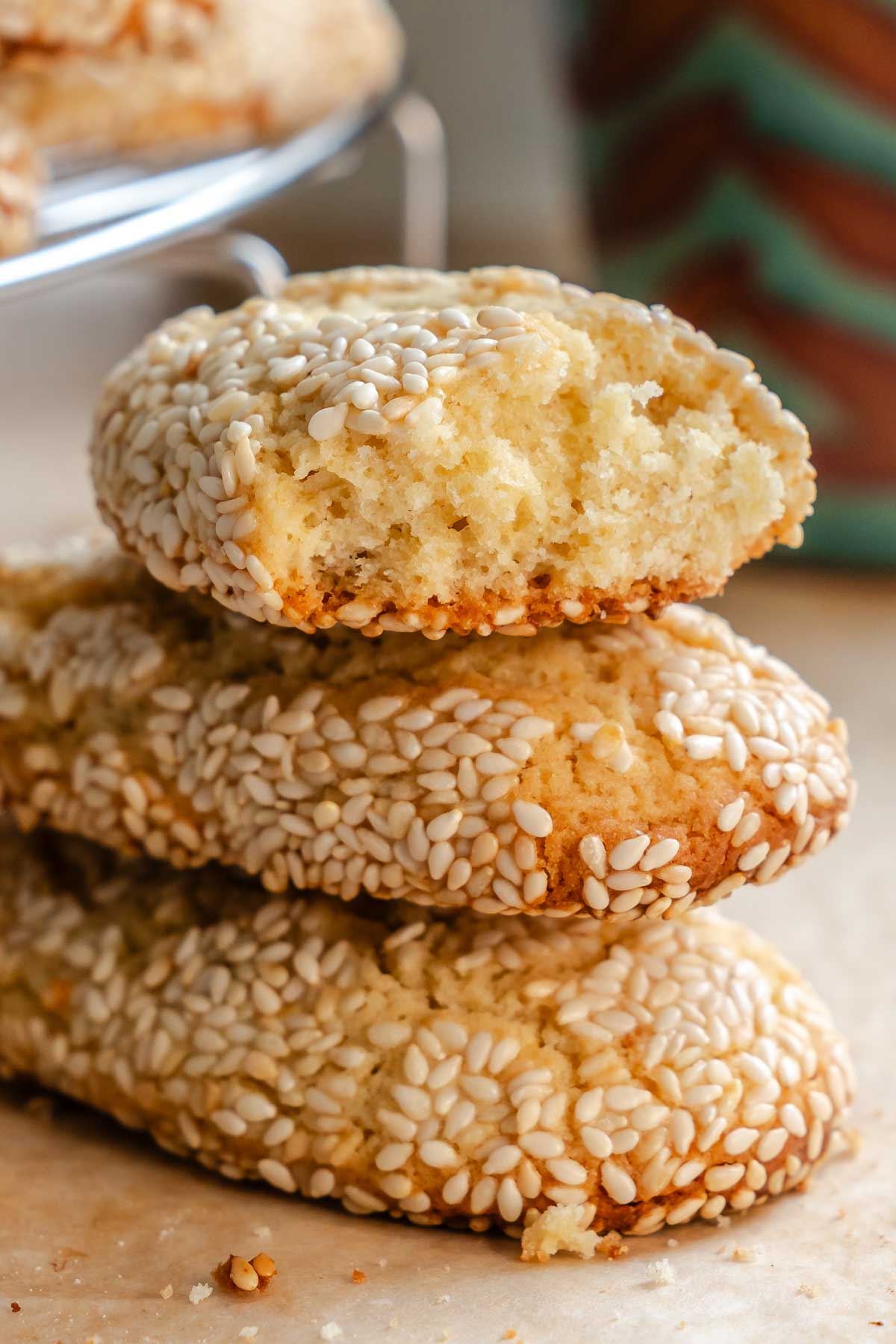 Stack of three Italian sesame seed cookies with the top cookie broken in half, revealing a soft, pale yellow, crumbly interior surrounded by a thick crust of white sesame seeds.