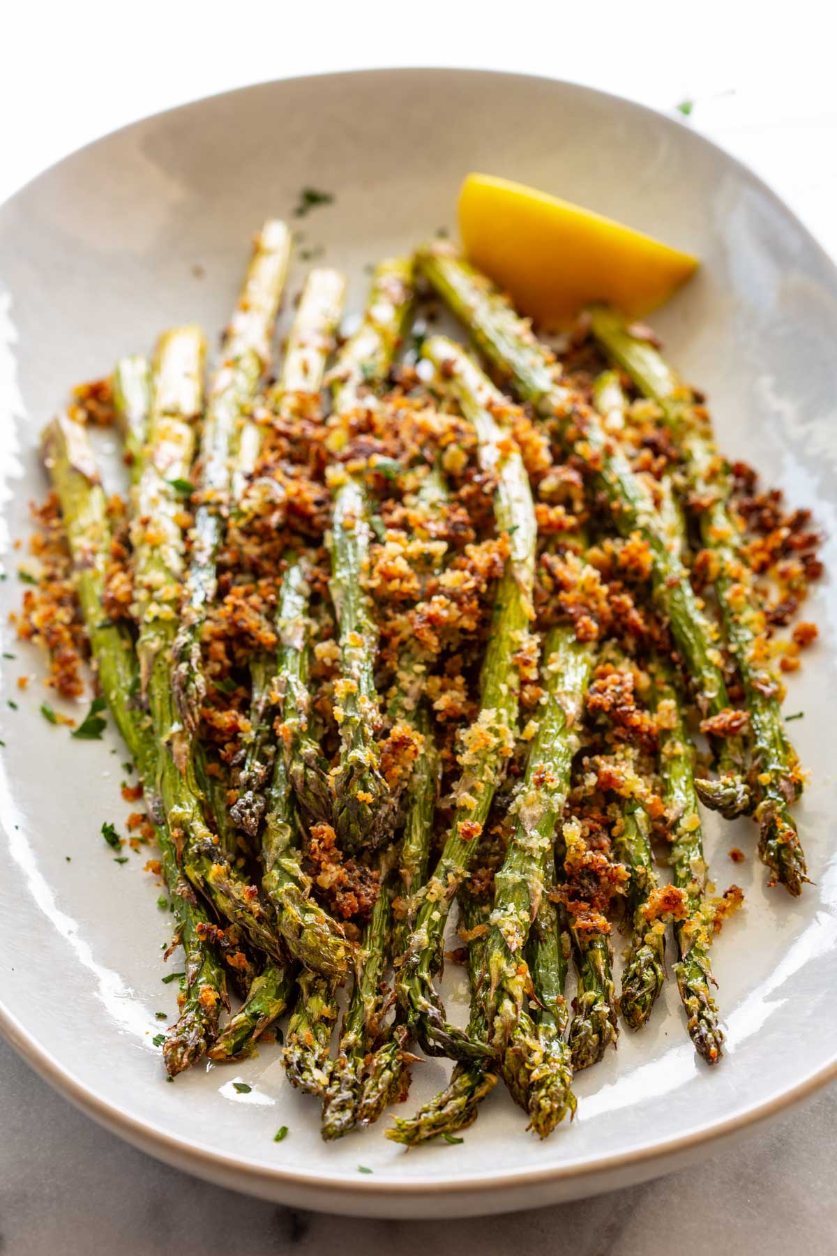 Roasted asparagus spears piled on a round white plate, topped with a crispy golden parmesan panko crust, garnished with a lemon wedge and a sprinkle of fresh parsley.