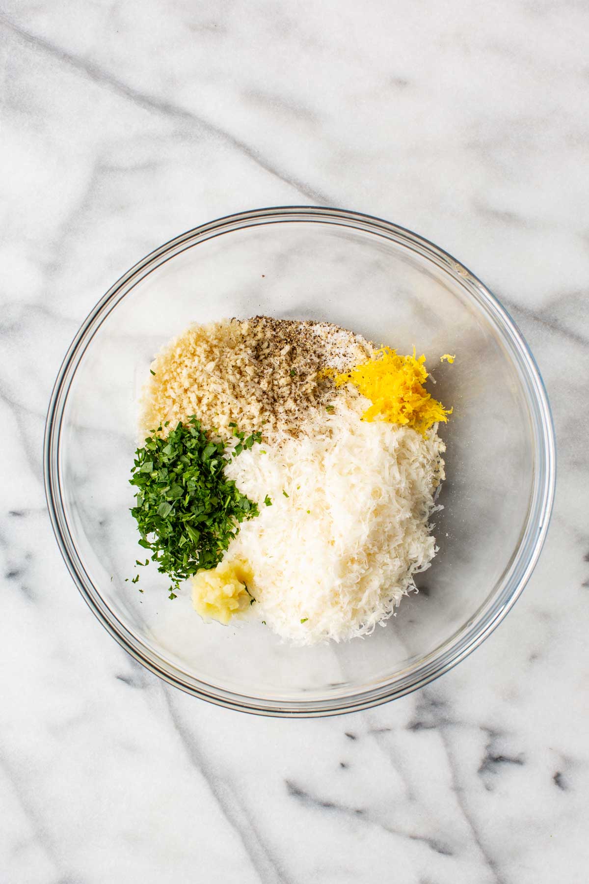 A glass bowl on a marble surface containing unmixed ingredients for a parmesan panko topping: shredded parmesan, panko breadcrumbs, chopped fresh parsley, lemon zest, minced garlic, and black pepper.