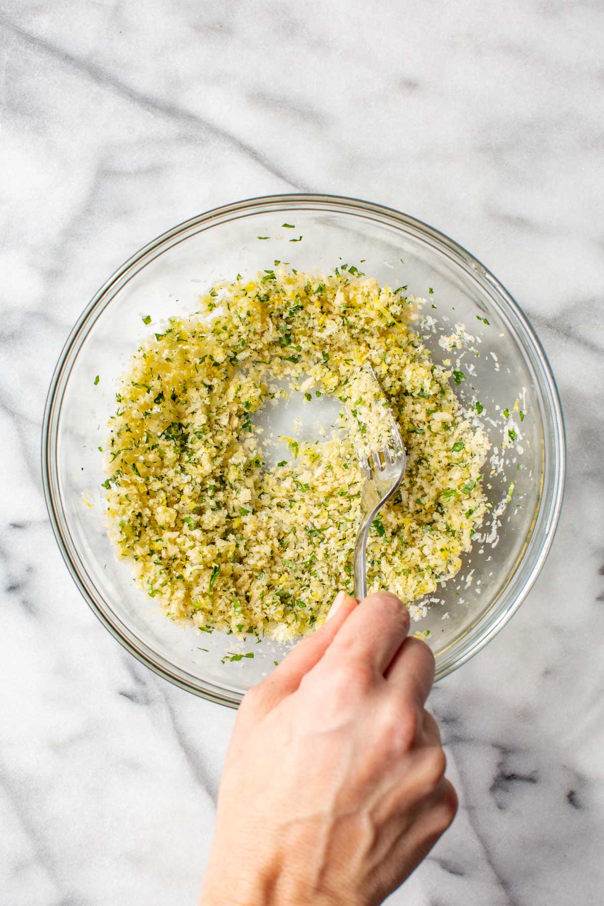 A hand using a fork to mix together the parmesan panko topping ingredients in a glass bowl on a marble surface, combining them into an even, crumbly mixture.