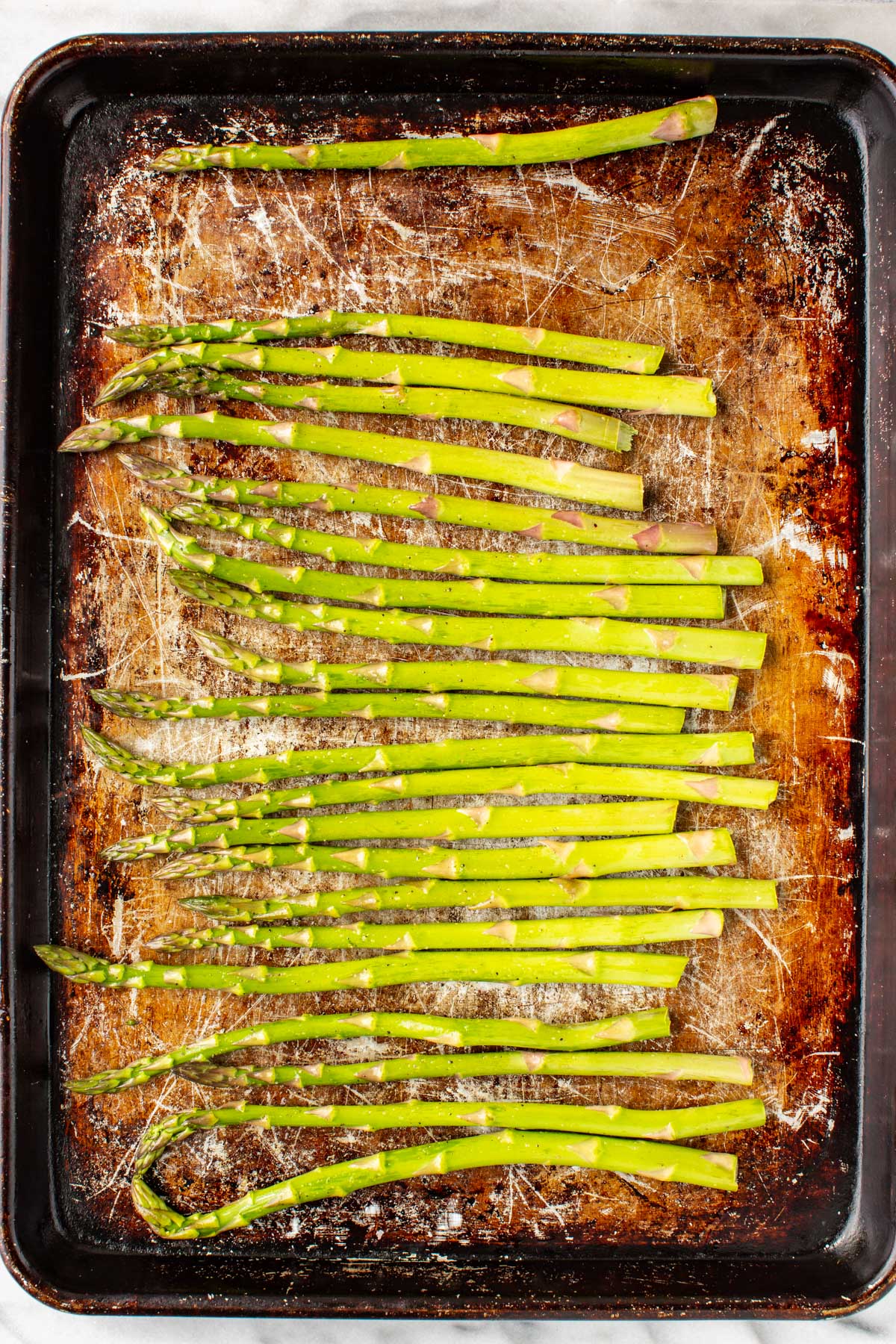 Raw asparagus spears arranged in a single layer on a well-seasoned dark baking sheet, trimmed and ready to be topped before roasting.