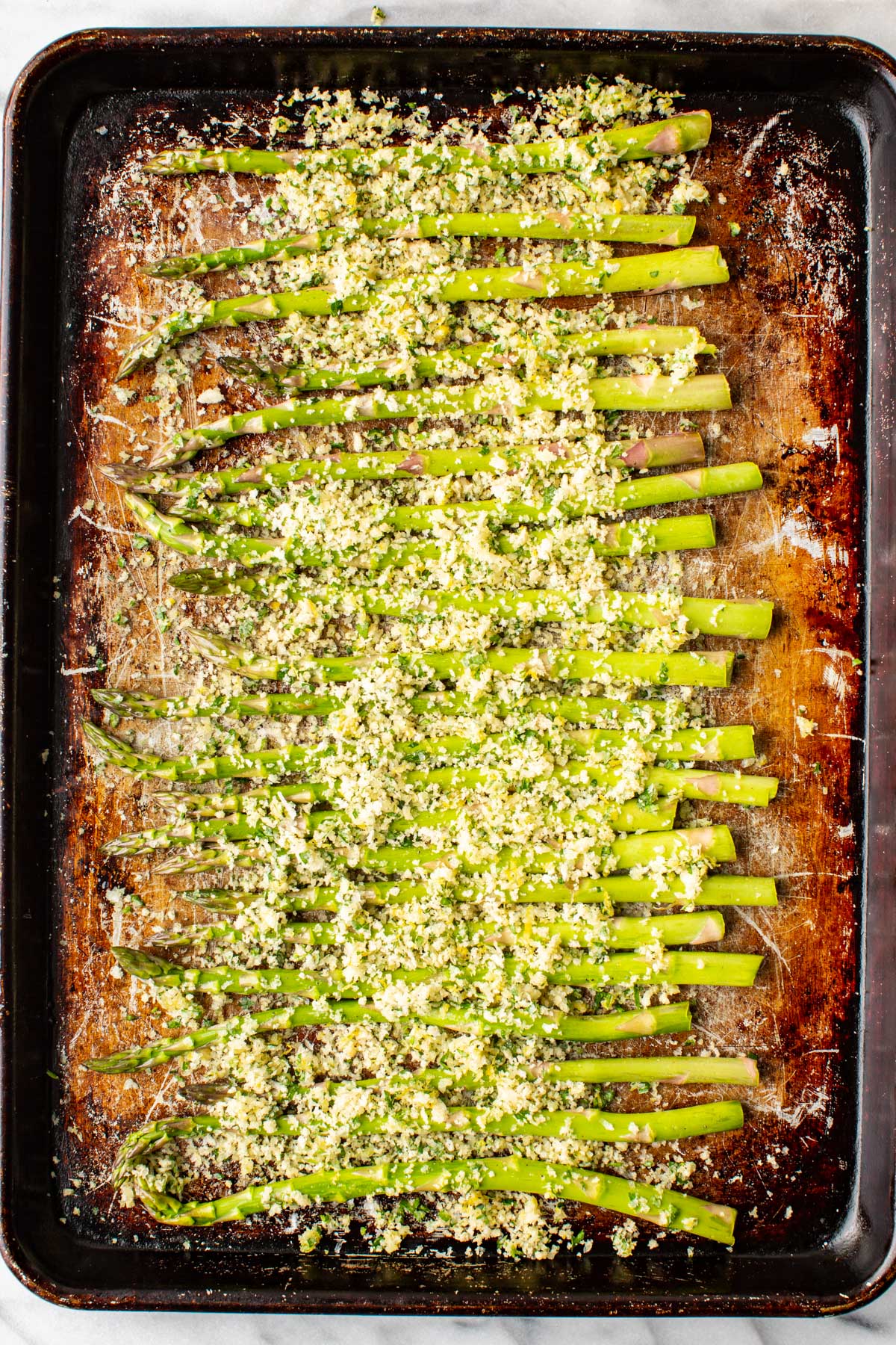Asparagus spears on a dark baking sheet, evenly coated with the parmesan panko mixture before going into the oven.