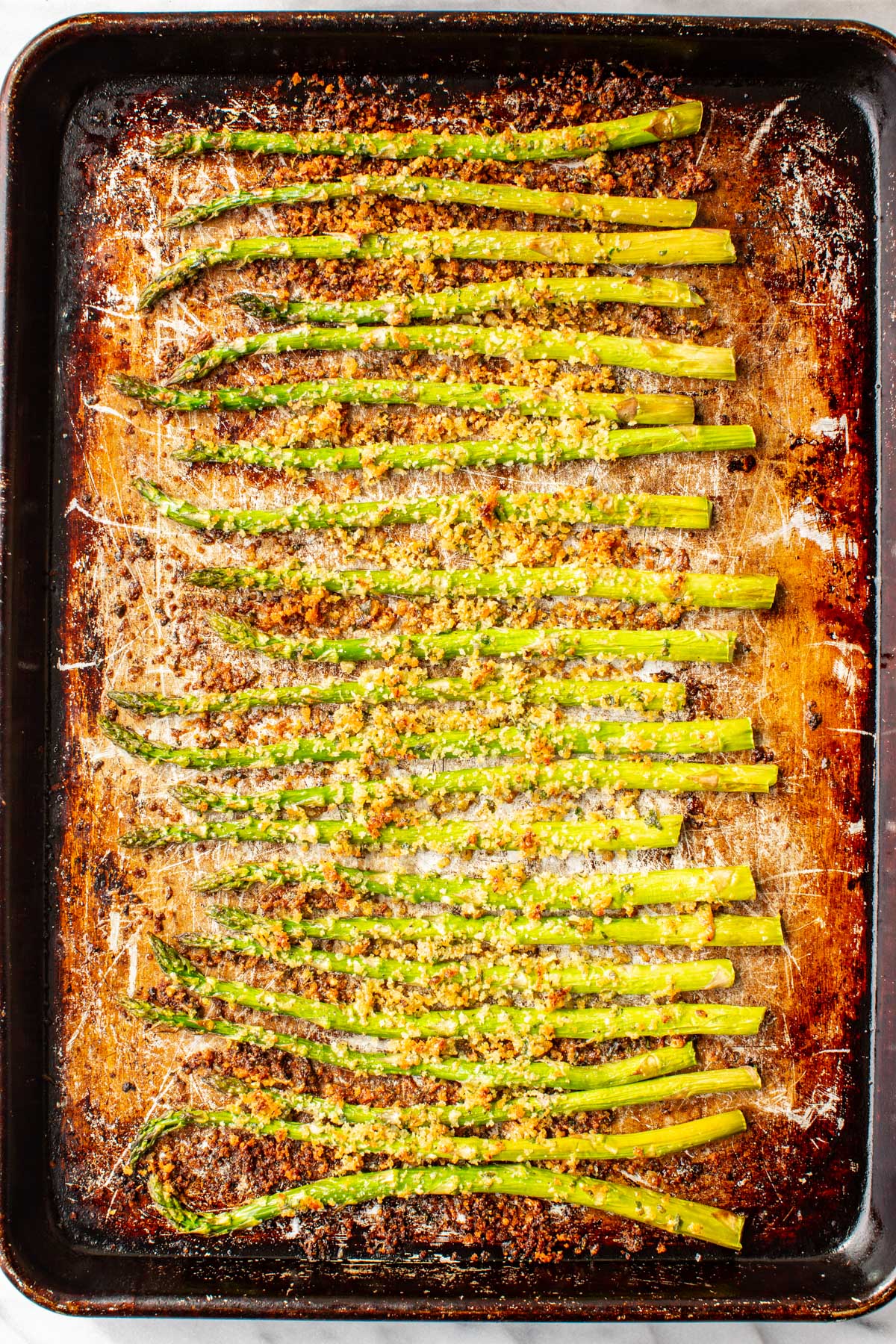 Asparagus spears fresh from the oven on a dark baking sheet, the parmesan panko topping deeply golden brown and crispy.