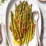 Overhead view of roasted asparagus with a golden parmesan panko crust arranged on a white oval serving platter, garnished with a lemon wedge, with small bowls of chopped parsley and lemon wedges alongside.