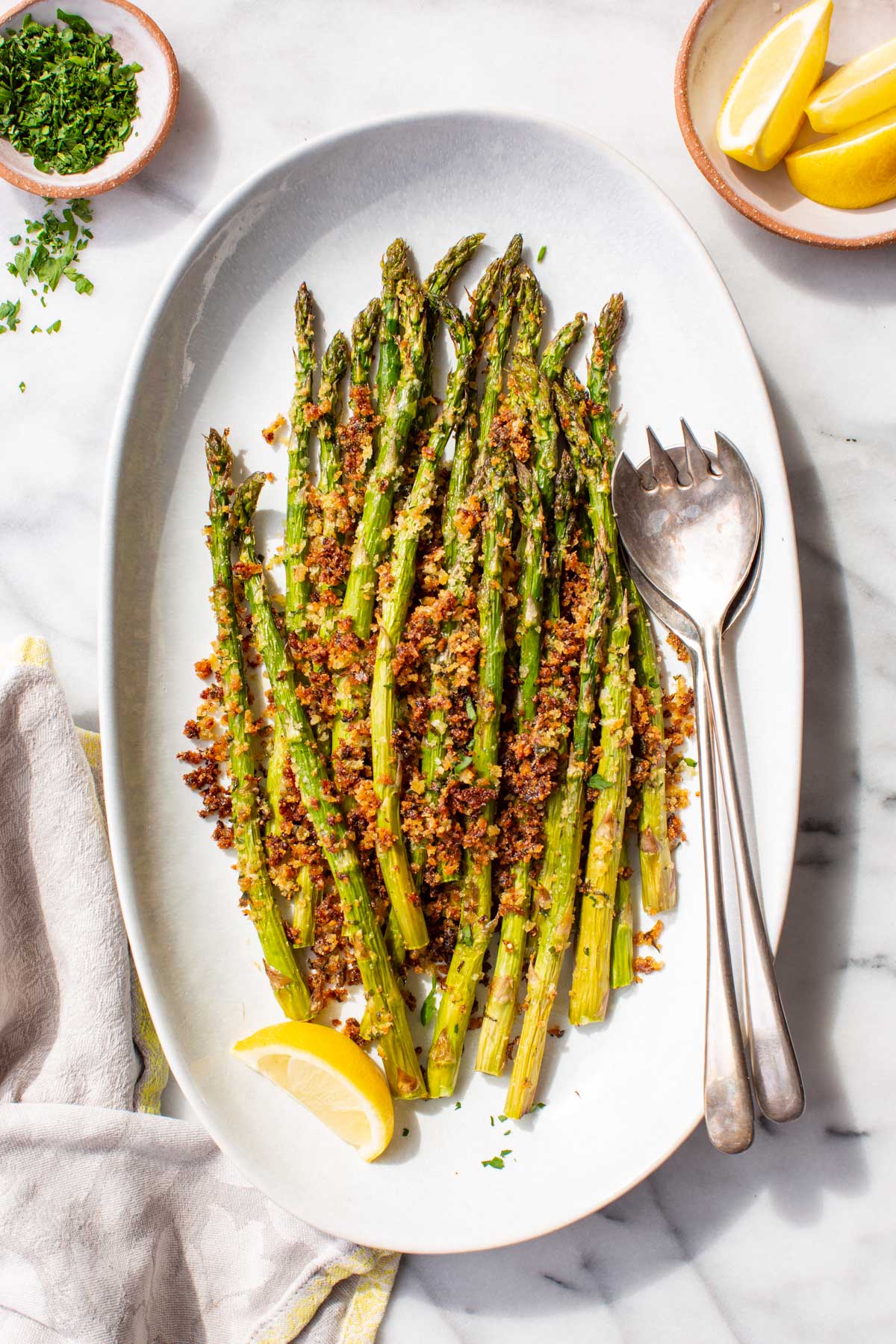 Overhead view of roasted asparagus with a golden parmesan panko crust arranged on a white oval serving platter, garnished with a lemon wedge, with small bowls of chopped parsley and lemon wedges alongside.