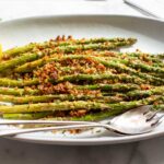 Side-angle view of crispy parmesan-crusted roasted asparagus on a white oval platter with a lemon wedge and serving utensils, a block of parmesan and fresh herbs visible in the background.