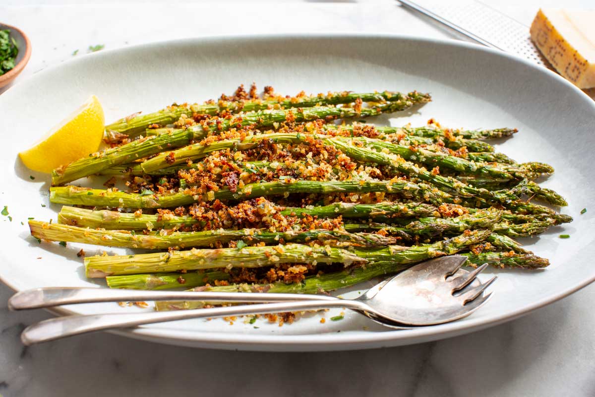 Side-angle view of crispy parmesan-crusted roasted asparagus on a white oval platter with a lemon wedge and serving utensils, a block of parmesan and fresh herbs visible in the background.