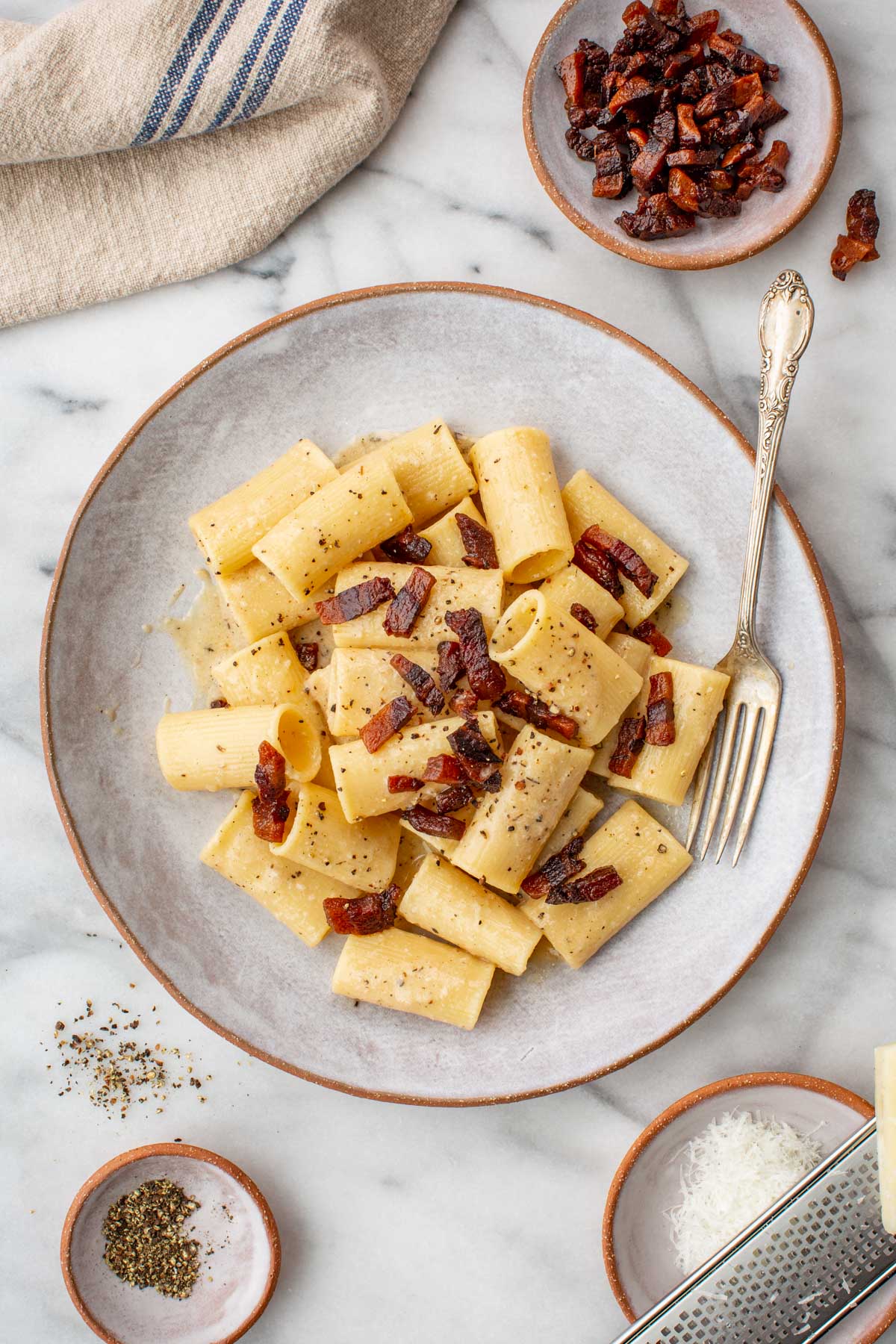 Rigatoni pasta tossed in a glossy pecorino cheese sauce and topped with crispy guanciale and cracked black pepper, served in a shallow bowl on a marble surface.