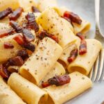 Tight close-up of rigatoni coated in creamy pecorino sauce with crisp browned guanciale pieces nestled in the ridges and finished with black pepper.