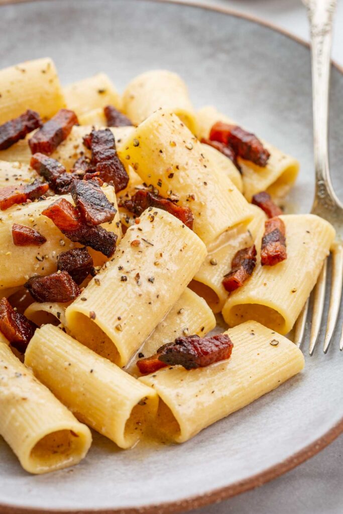 Tight close-up of rigatoni coated in creamy pecorino sauce with crisp browned guanciale pieces nestled in the ridges and finished with black pepper.
