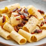Rigatoni pasta in a creamy cheese sauce with scattered crispy guanciale, shown from a low angle highlighting the texture of the pasta and sauce.