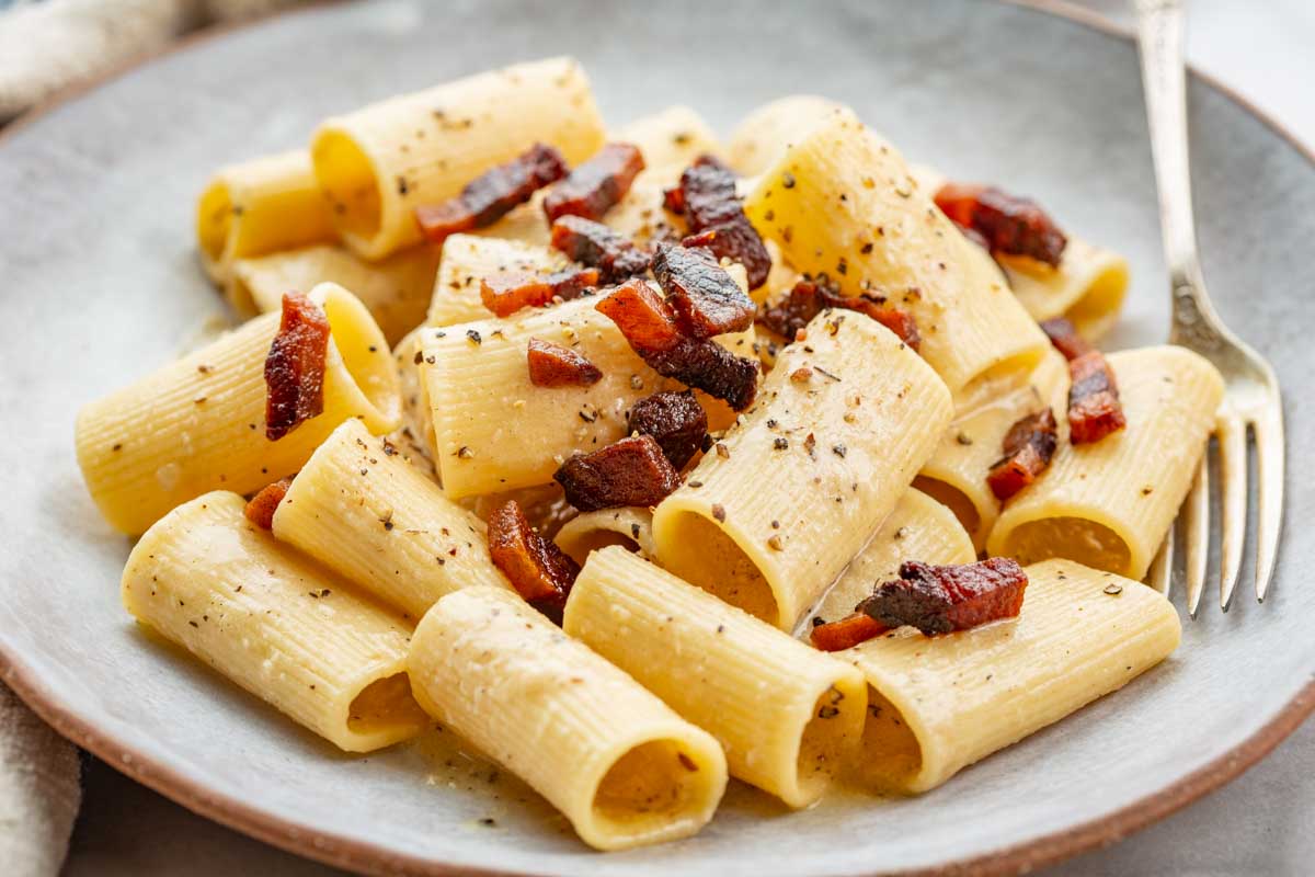 Rigatoni pasta in a creamy cheese sauce with scattered crispy guanciale, shown from a low angle highlighting the texture of the pasta and sauce.