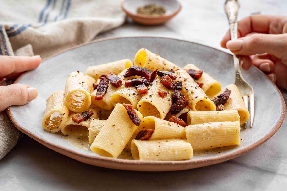 Hands holding a fork over a bowl of rigatoni tossed in pecorino sauce with crispy guanciale and black pepper, ready to eat.