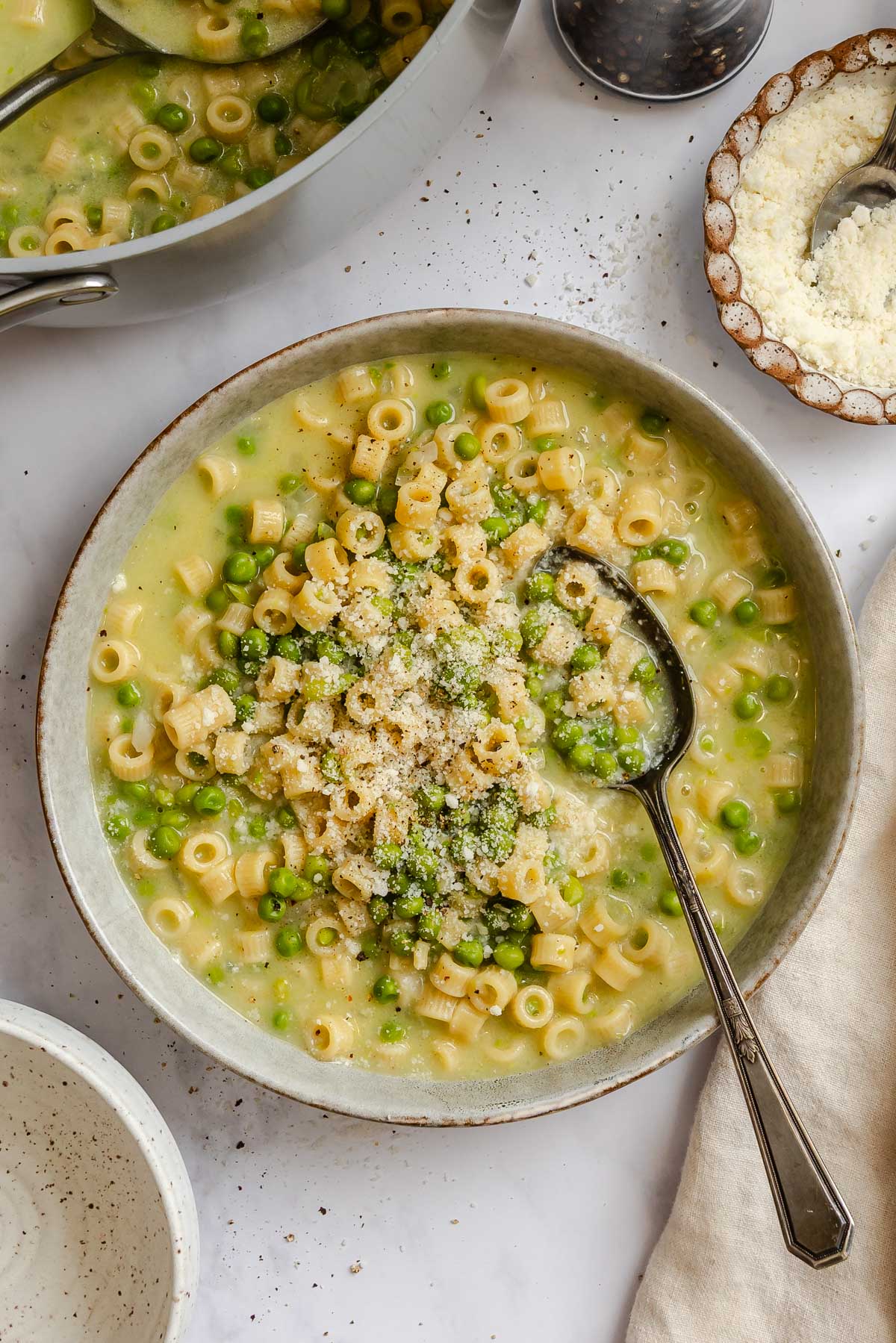 Overhead view of a bowl of pasta e piselli topped with grated Parmesan cheese, with the pot and extra cheese on the side.