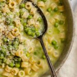 Close up of pasta e piselli in a shallow bowl with a spoon scooping ditalini, peas and broth.
