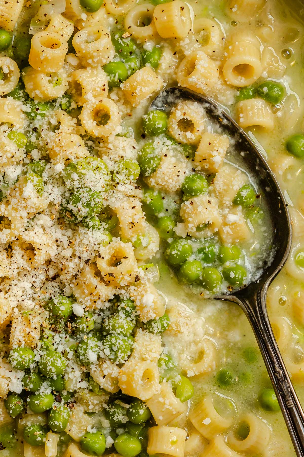 Extreme close up of ditalini pasta and green peas in broth topped with grated Parmesan and cracked black pepper.