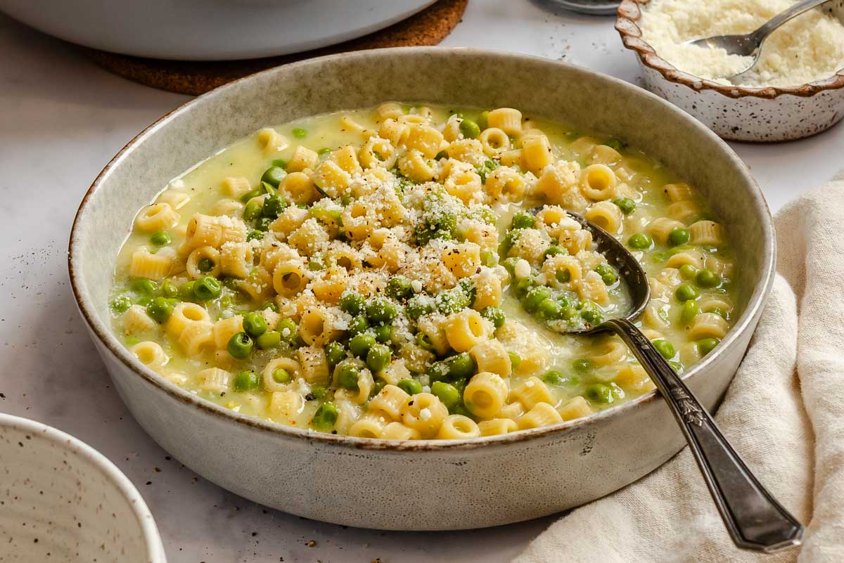 Wide view of a bowl of pasta e piselli topped with grated Parmesan, served with a spoon and extra cheese on the side.