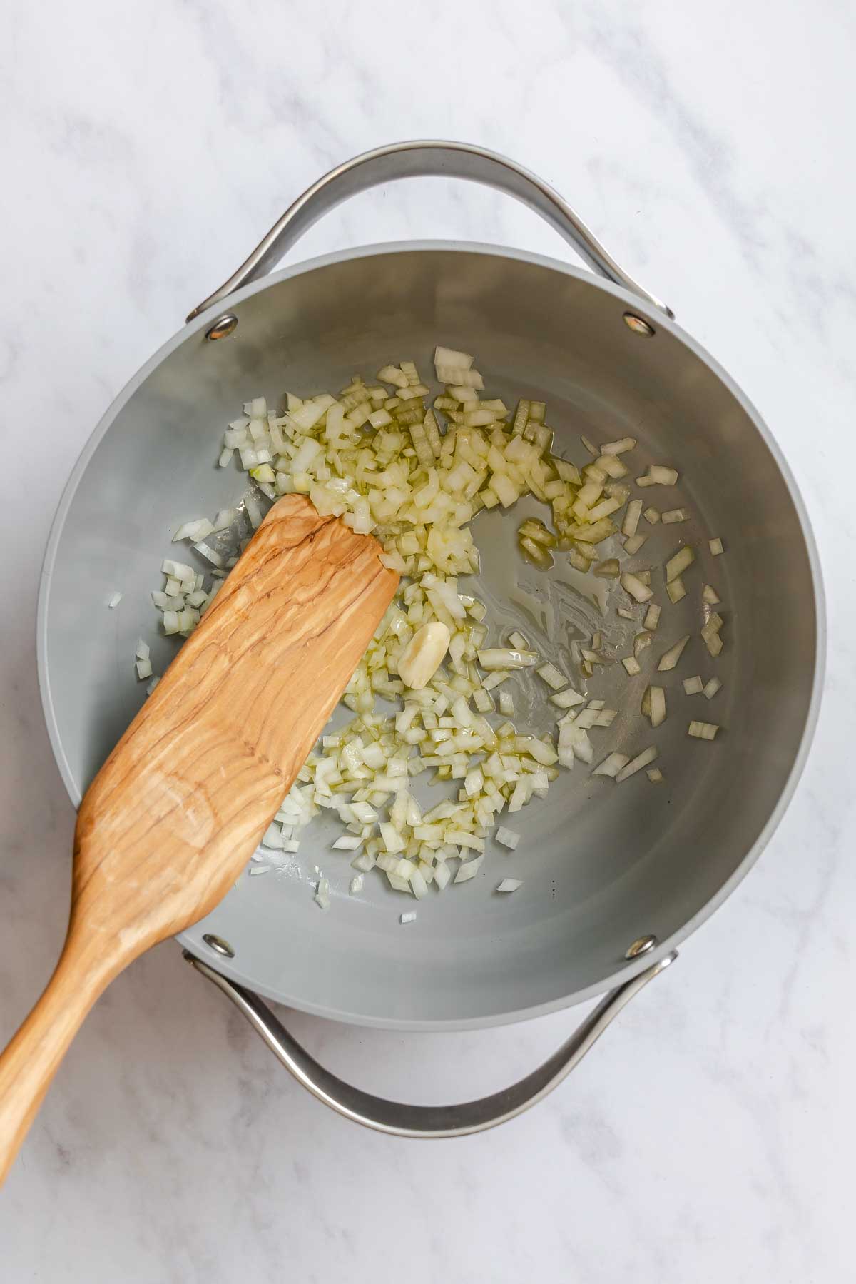 Onions and garlic being sauteed in a gray pot with with a wooden spoon.