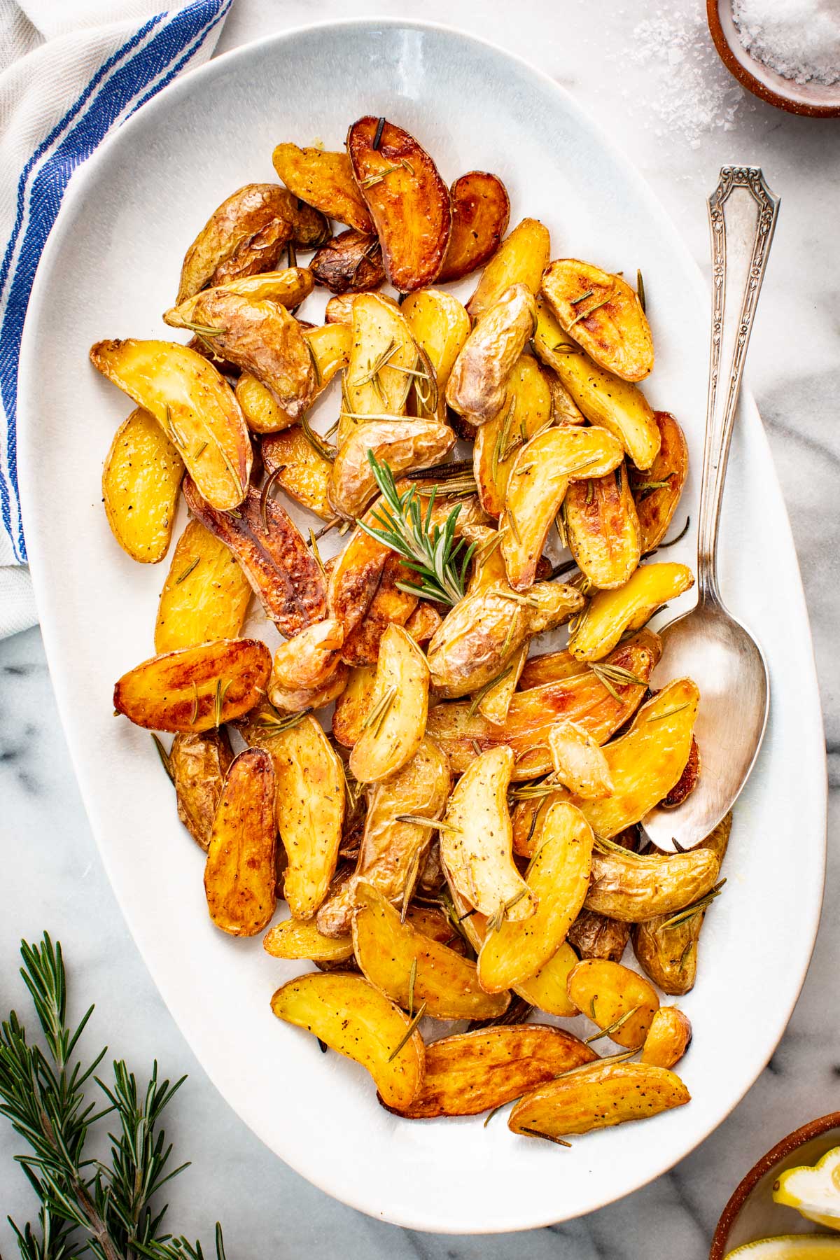 Roasted fingerling potatoes tossed with rosemary and garlic, served on a white platter with a spoon and coarse salt on the side.