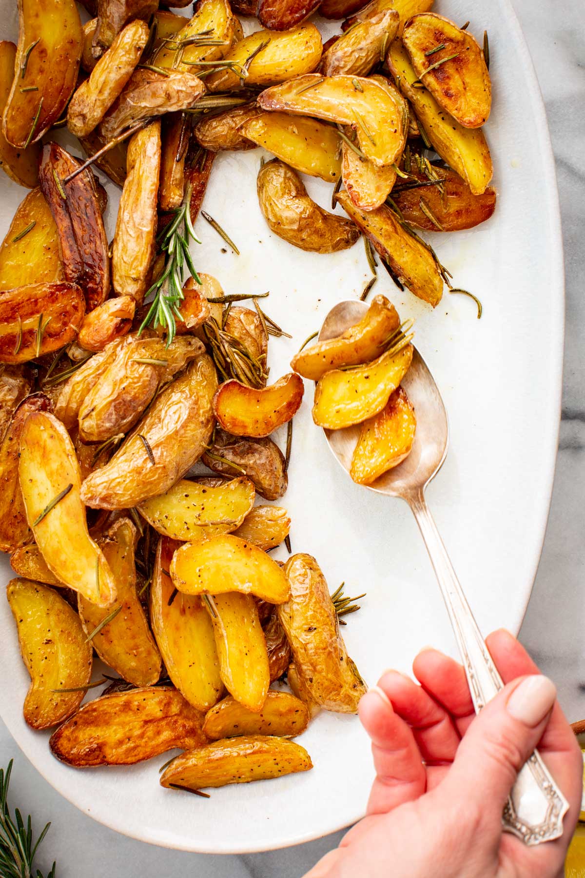 Hand scooping crispy roasted fingerling potatoes with rosemary from a white serving platter using a spoon.