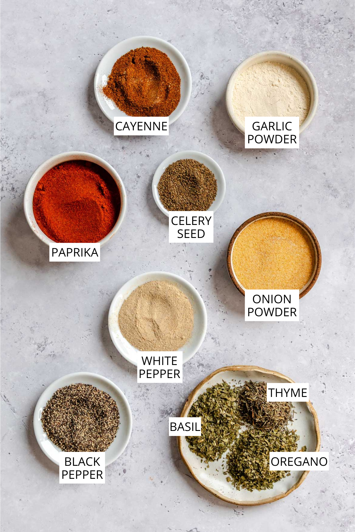 Flat lay of individual Cajun seasoning ingredients in small bowls on a gray surface, labeled: cayenne, garlic powder, paprika, celery seed, onion powder, white pepper, black pepper, basil, thyme, and oregano.