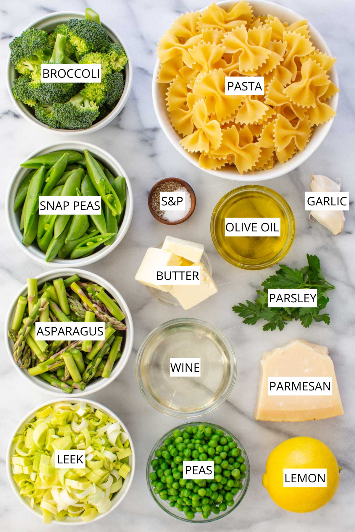 Flat lay of pasta primavera ingredients on a marble surface, labeled: farfalle pasta, broccoli, snap peas, asparagus, leek, peas, garlic, olive oil, butter, white wine, parmesan, parsley, lemon, and salt & pepper.