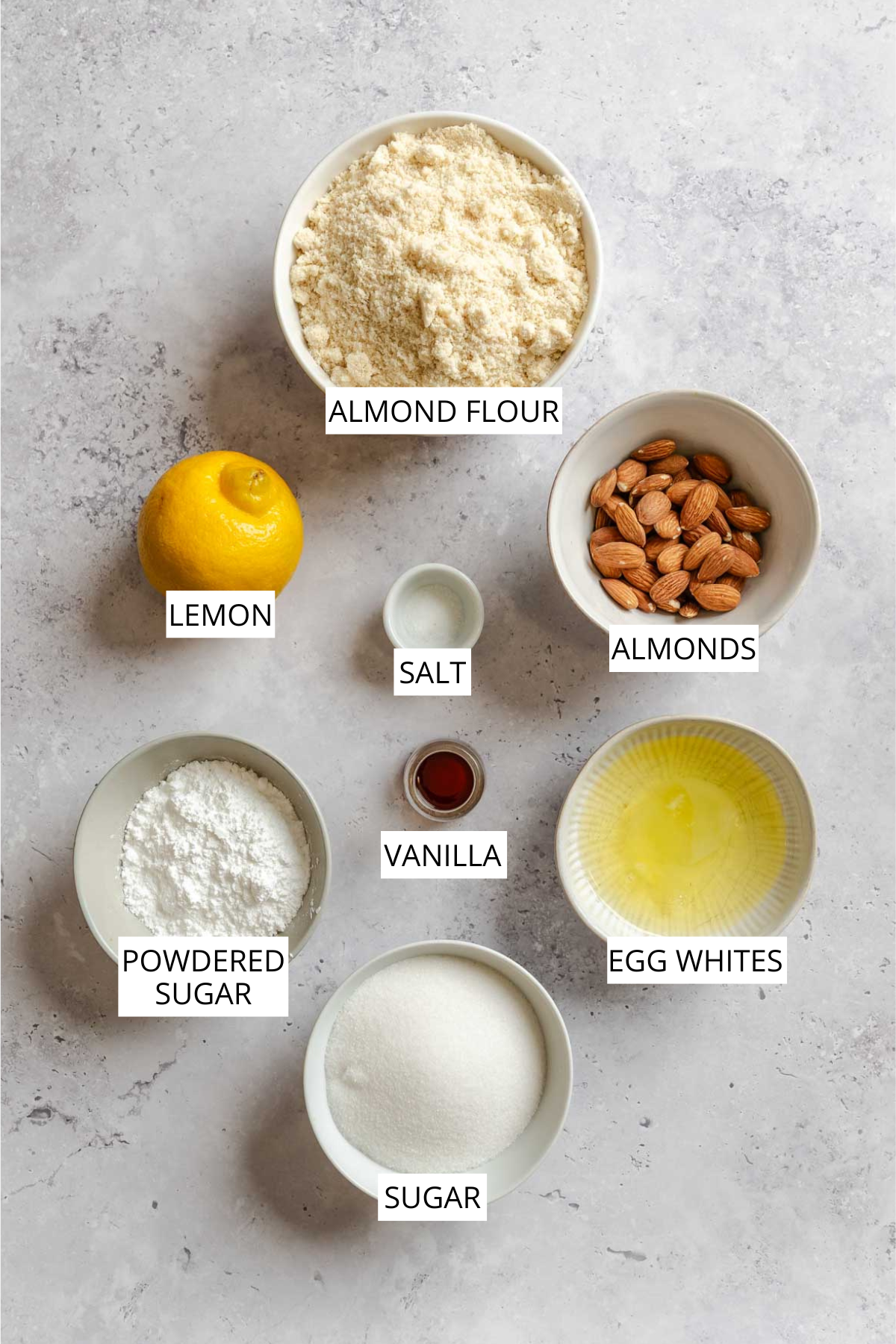 Overhead flat-lay of almond cookies ingredients including almond flour, lemon, almonds, salt, vanilla, sugar and egg whites.