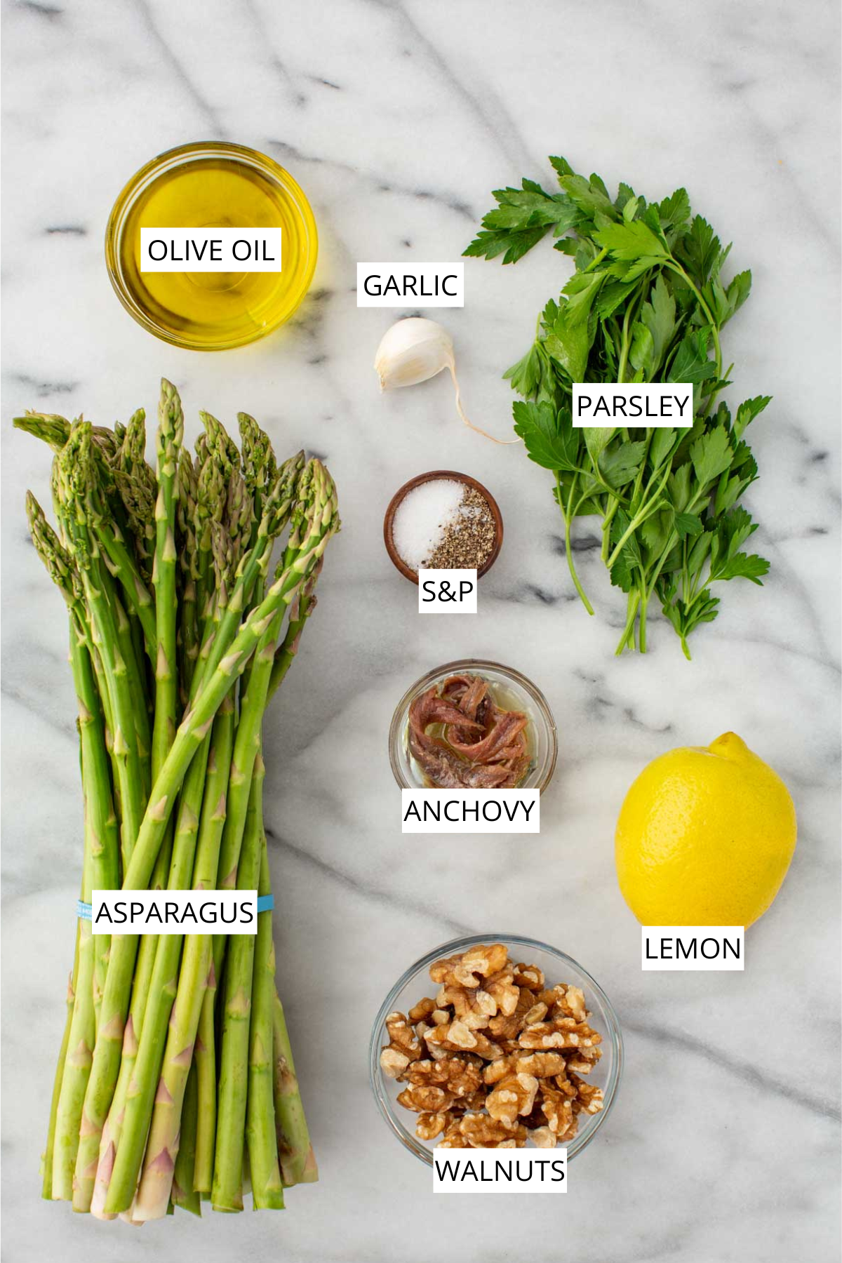 Overhead flat lay of labeled ingredients for grilled asparagus on a marble surface: a bunch of fresh asparagus, a small bowl of olive oil, a garlic clove, fresh parsley, a small bowl of salt and pepper, a small glass bowl of anchovy fillets, a whole lemon, and a glass bowl of walnuts.