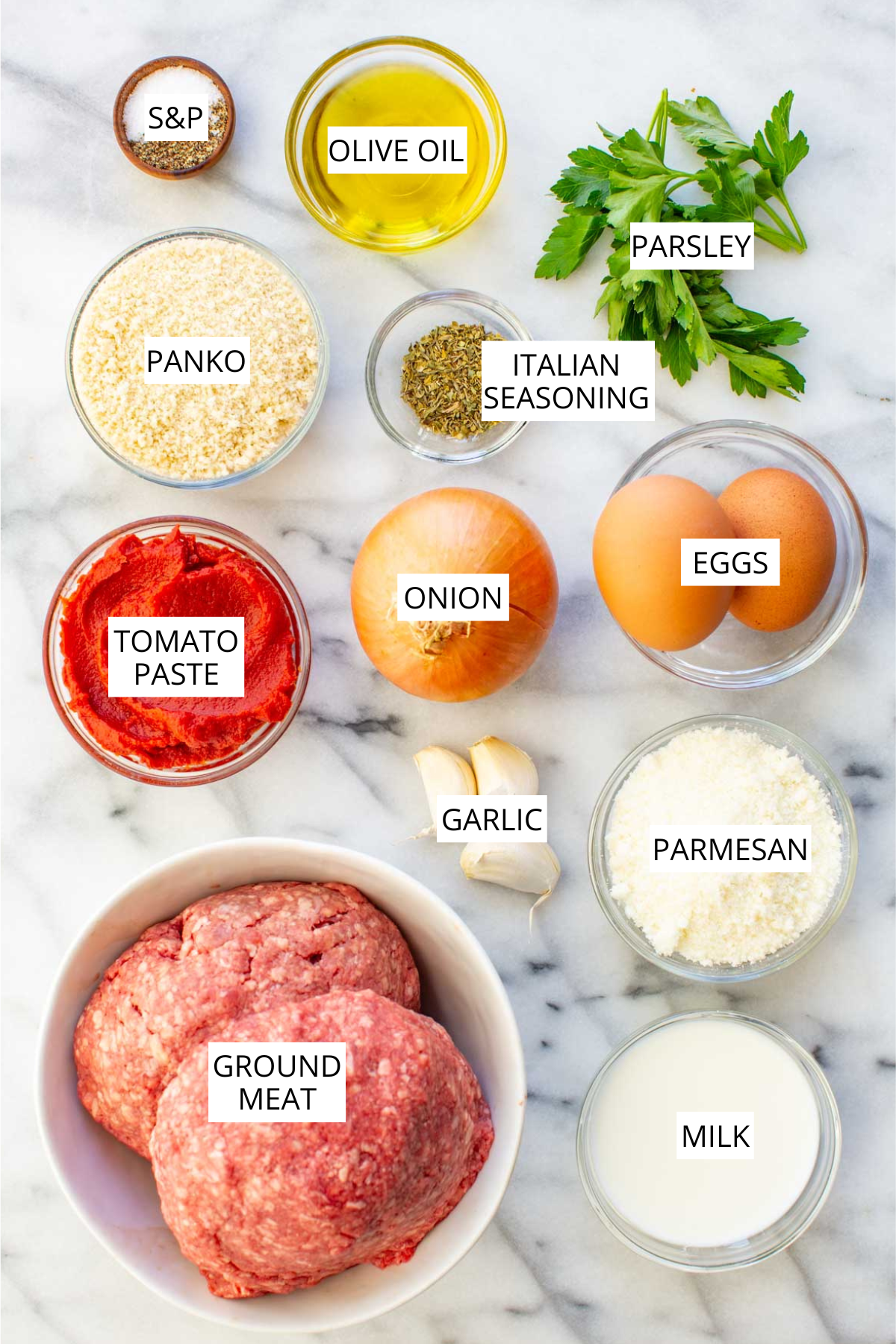 Flat lay of Italian meatloaf ingredients on a marble surface, labeled: ground meat, eggs, milk, parmesan, garlic, onion, tomato paste, panko breadcrumbs, Italian seasoning, parsley, olive oil, and salt & pepper.