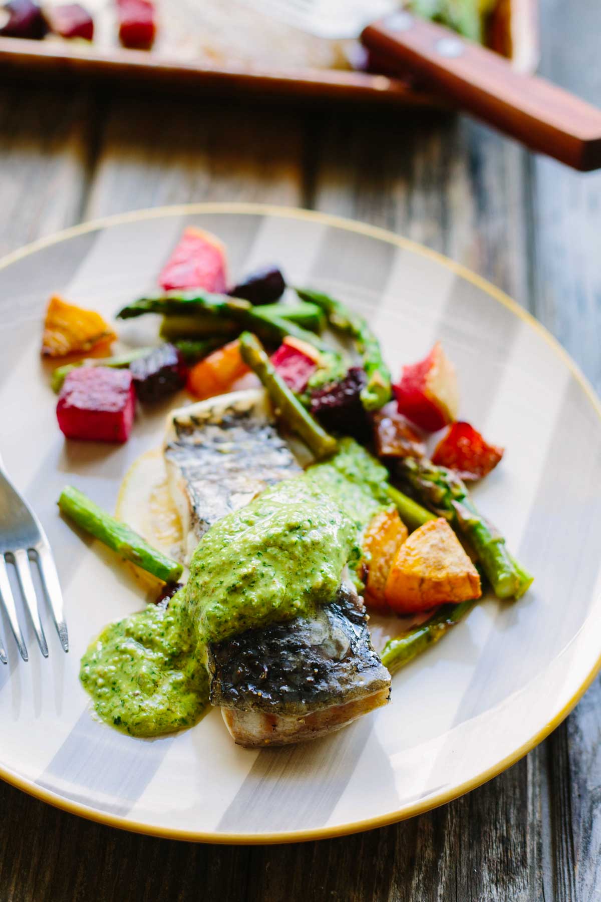 A plate of baked fish fillet topped with vibrant green arugula pesto, served alongside roasted asparagus and colorful cubed beets in red, golden, and purple hues, on a striped white and gold-rimmed plate with a fork.
