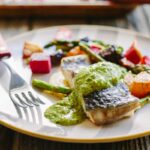 A close-up of a baked fish fillet drizzled with bright green arugula pesto on a gold-rimmed plate, accompanied by roasted beets and asparagus, with a fork and steak knife resting on the plate.