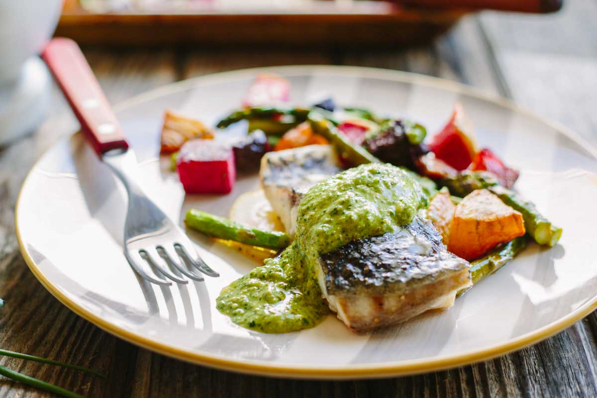 A close-up of a baked fish fillet drizzled with bright green arugula pesto on a gold-rimmed plate, accompanied by roasted beets and asparagus, with a fork and steak knife resting on the plate.