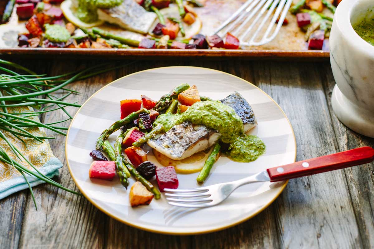 A full plate view of baked fish with arugula pesto over roasted spring vegetables including asparagus and tri-colored beets on a gold-rimmed striped plate, with a sheet pan of the full batch visible in the background.