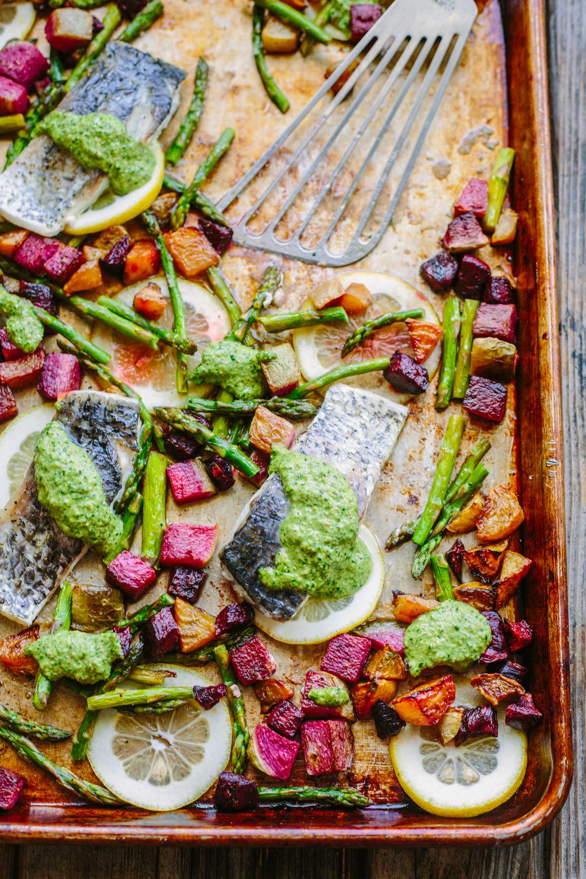 An overhead close-up of a sheet pan filled with baked fish fillets on lemon slices, dolloped with green arugula pesto, surrounded by roasted asparagus and colorful cubed beets, with a metal spatula resting in the pan.