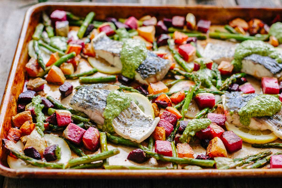 A wide shot of a full sheet pan of baked fish fillets topped with arugula pesto, nestled among roasted asparagus spears and vibrant red, golden, and purple beet chunks on lemon slices.