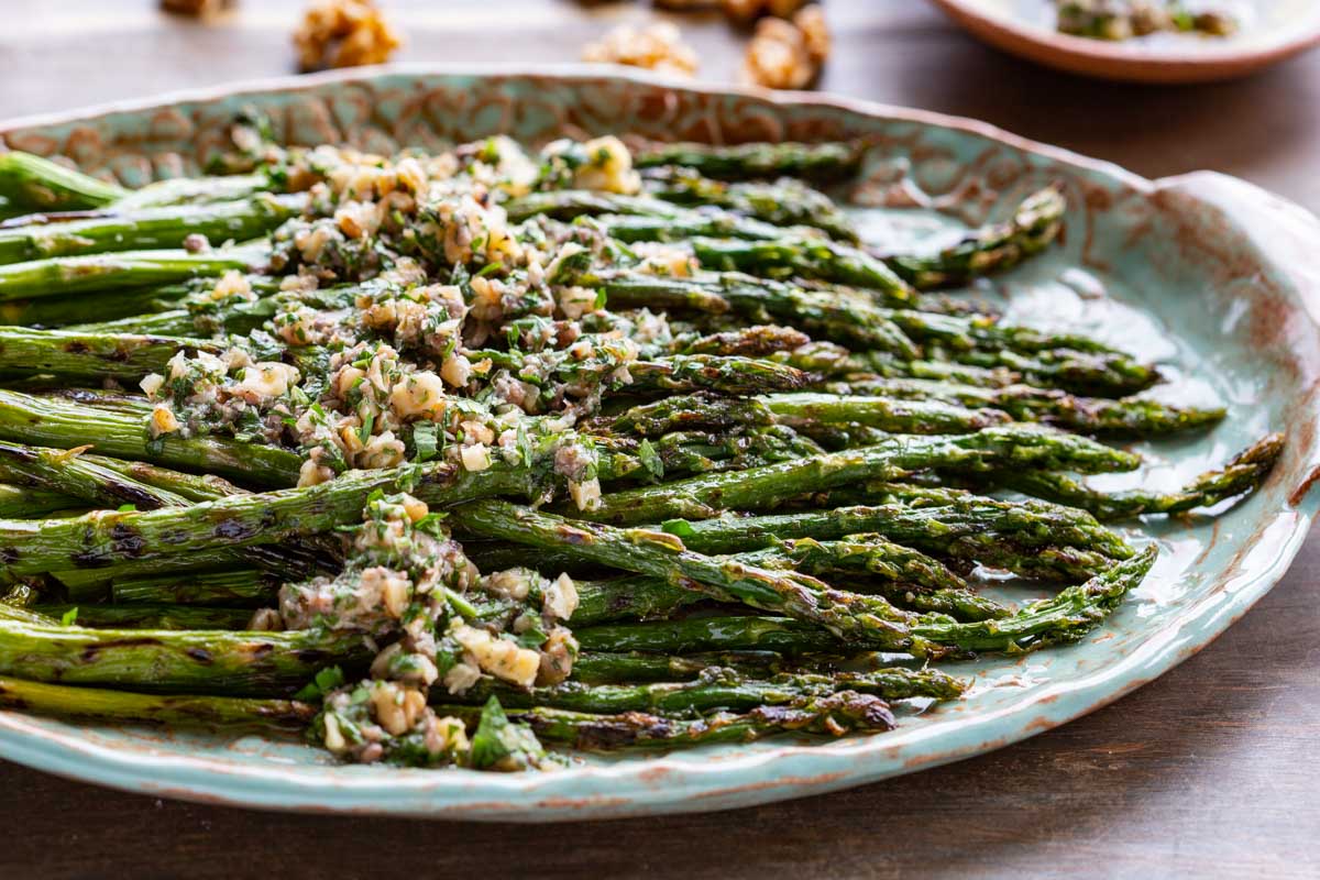 A side-angle view of charred grilled asparagus with walnut herb sauce on a teal ceramic platter, with whole walnuts blurred in the background.