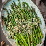 Overhead view of grilled asparagus with walnut herb sauce on a decorative teal platter, surrounded by whole walnuts, a garlic bulb, a halved lemon, and a small bowl of sauce on a dark wood surface.