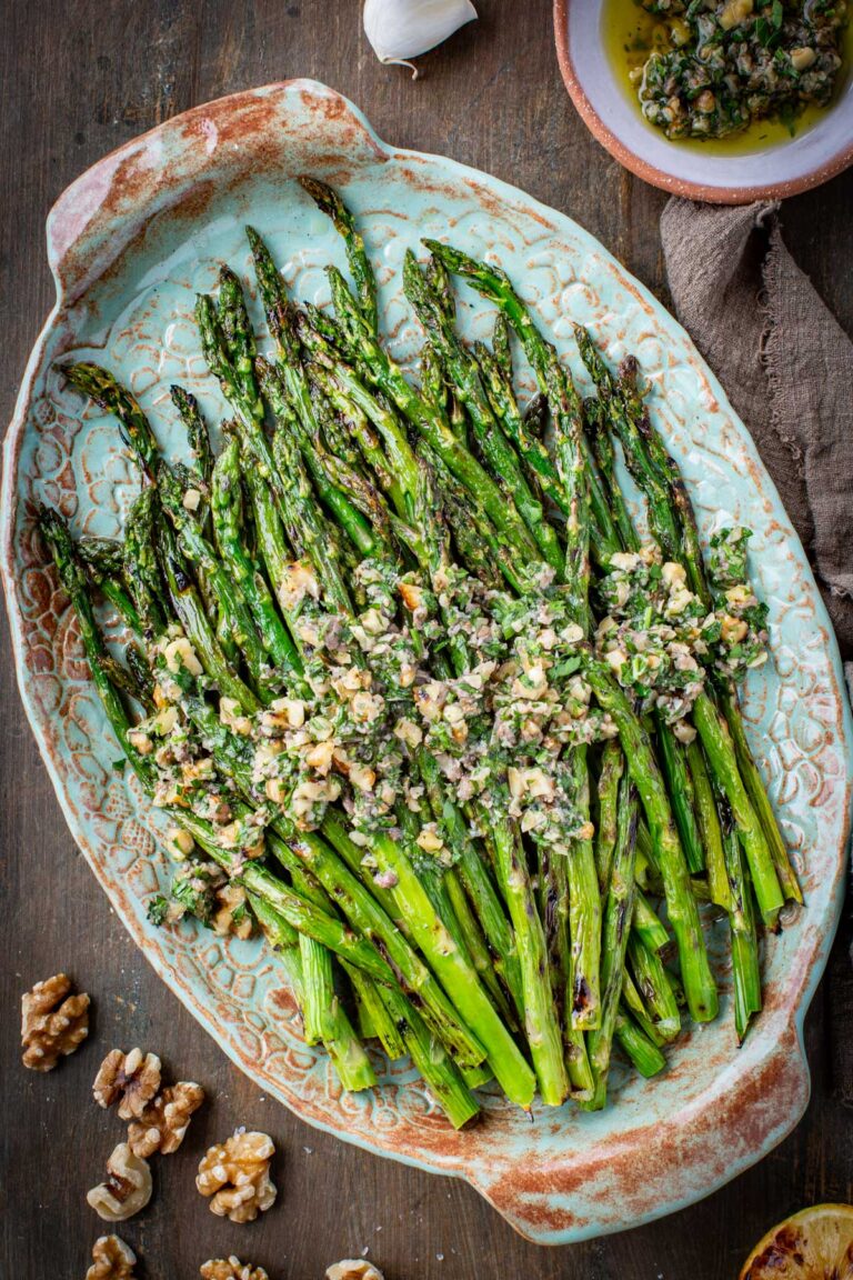 Overhead view of grilled asparagus with walnut herb sauce on a decorative teal platter, surrounded by whole walnuts, a garlic bulb, a halved lemon, and a small bowl of sauce on a dark wood surface.