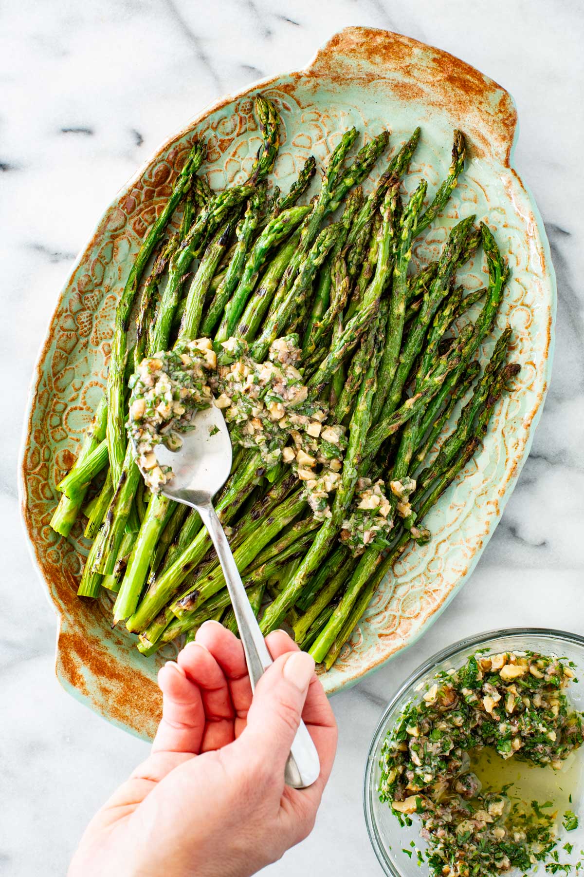 A hand spooning walnut herb sauce over grilled asparagus spears arranged on a teal and rust ceramic oval platter, with a small glass bowl of the sauce visible in the corner, set on a marble surface.