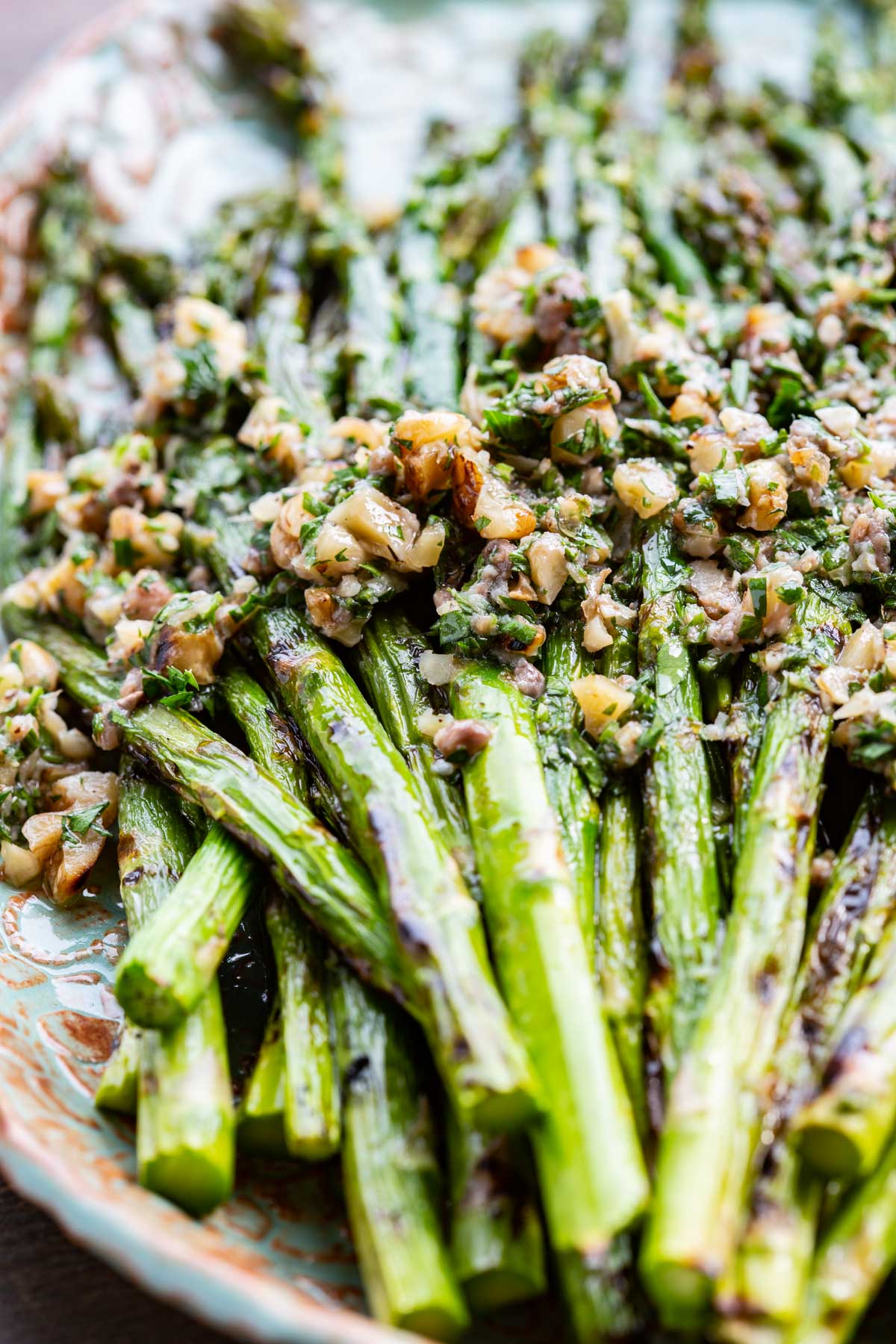 Close-up of grilled asparagus spears topped with a chunky walnut, garlic, and fresh herb sauce on a decorative ceramic platter.