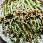 Grilled asparagus spears glistening with olive oil and topped with a walnut herb mixture, served on a teal ceramic platter with a scalloped edge.