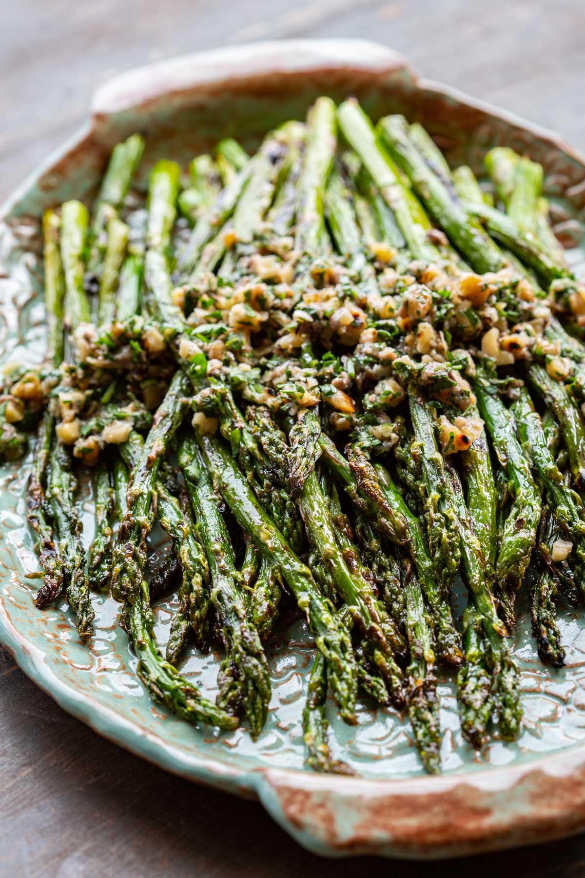 Grilled asparagus spears glistening with olive oil and topped with a walnut herb mixture, served on a teal ceramic platter with a scalloped edge.