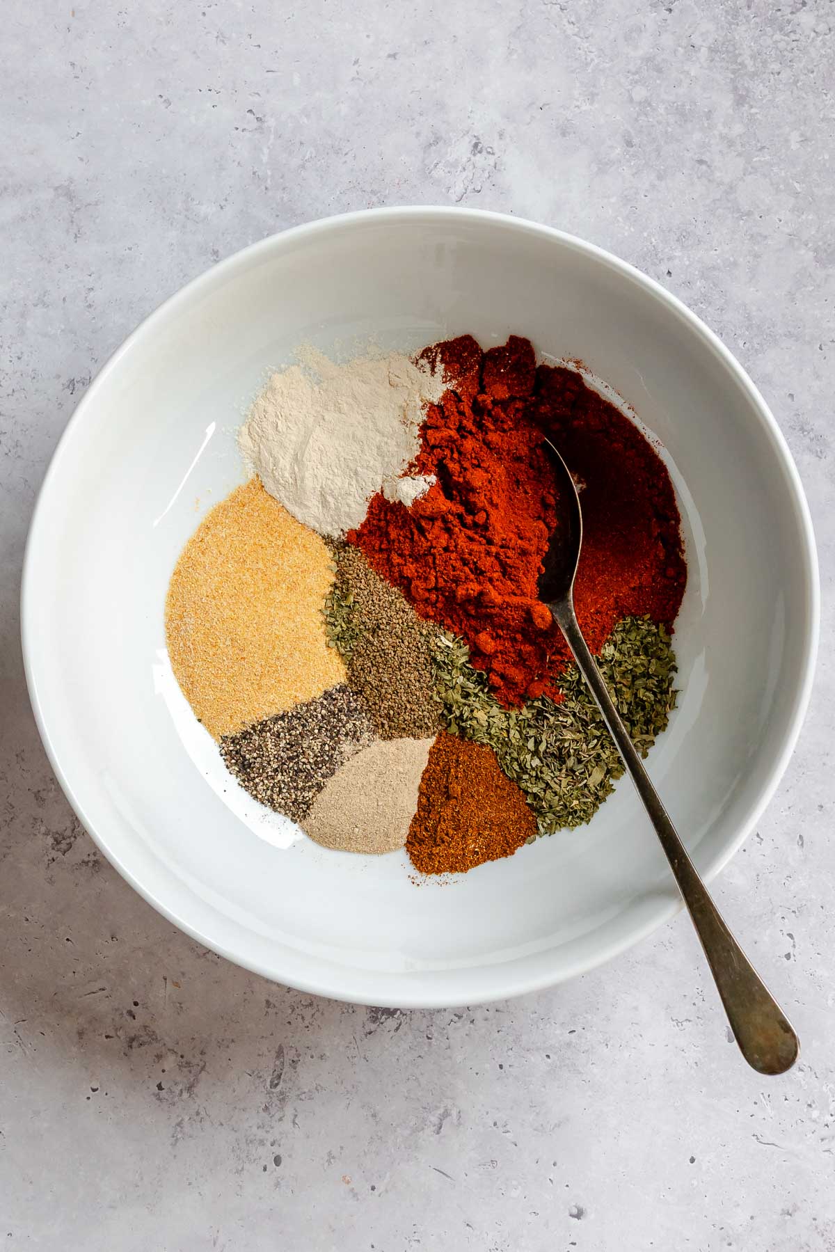 Overhead view of a large white bowl containing unmixed Cajun seasoning spices arranged in separate mounds - including paprika, onion powder, garlic powder, black pepper, herbs, and cayenne, with a vintage spoon resting inside.