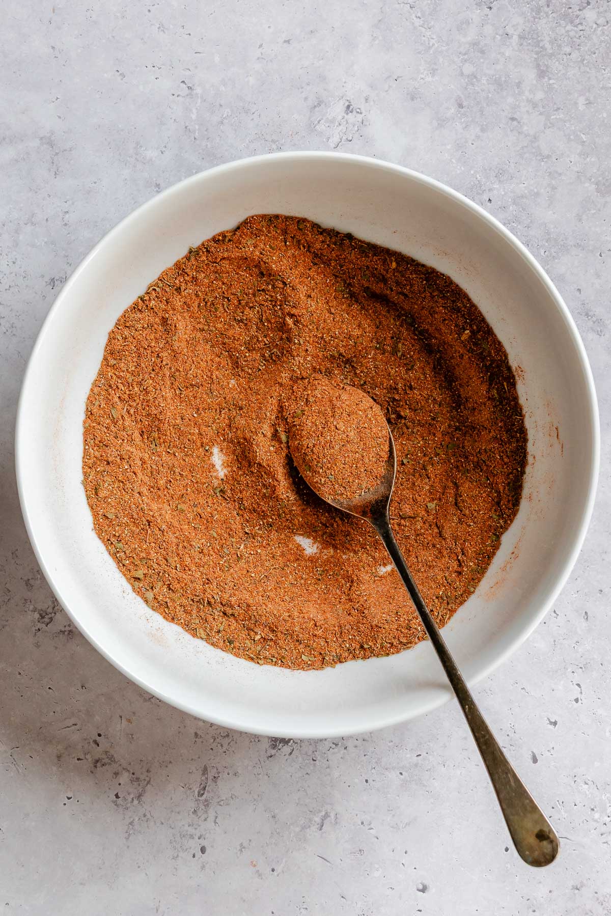 Overhead view of a large white bowl filled with fully blended Cajun seasoning, showing a uniform reddish-orange powder with flecks of dried herbs, with a spoon holding a heaping portion.