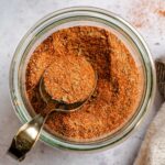 Close-up overhead view of homemade Cajun seasoning in a glass Weck jar, with a vintage measuring spoon scooping the vibrant reddish-orange spice blend.