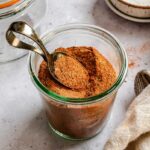 Side-angle view of a glass jar filled to the brim with homemade Cajun seasoning, a vintage spoon resting on top, with additional empty jars and a linen napkin in the background.