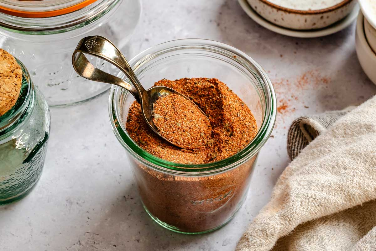Side-angle view of a glass jar filled to the brim with homemade Cajun seasoning, a vintage spoon resting on top, with additional empty jars and a linen napkin in the background.