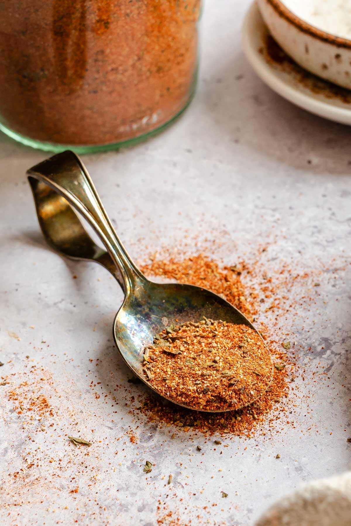 Close-up of a vintage spoon resting on a light stone surface, holding a small amount of Cajun seasoning with visible herb flecks, with a filled glass jar slightly blurred in the background.